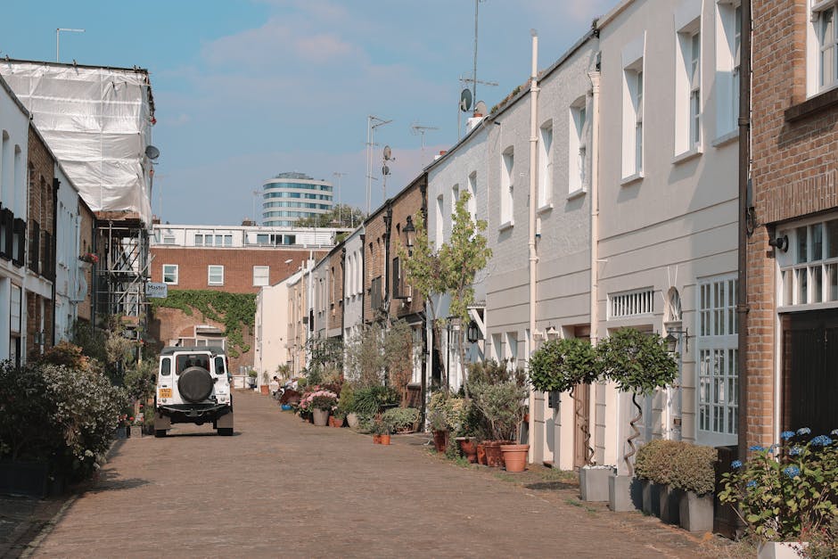 A narrow residential garden street with brick and white-painted townhouse facades, neatly lined with potted plants and small trees along the front of the houses. The paving is composed of cobblestones