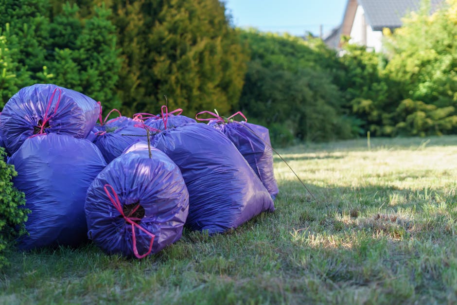 A row of purple garden waste bags filled with plant debris, tied with pink string, stacked on a grassy lawn next to a hedge with lush green foliage. In the background, there are trees, shrubs, and par